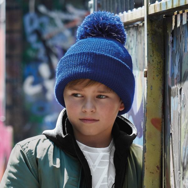 Bimbo con giacca e berretto con pom pom blu royal appoggiato a parete in strada