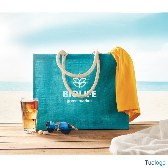 Shopper in juta azzurra con logo bianco e con manici corti bianchi, su tavolo di legno con dietro mare